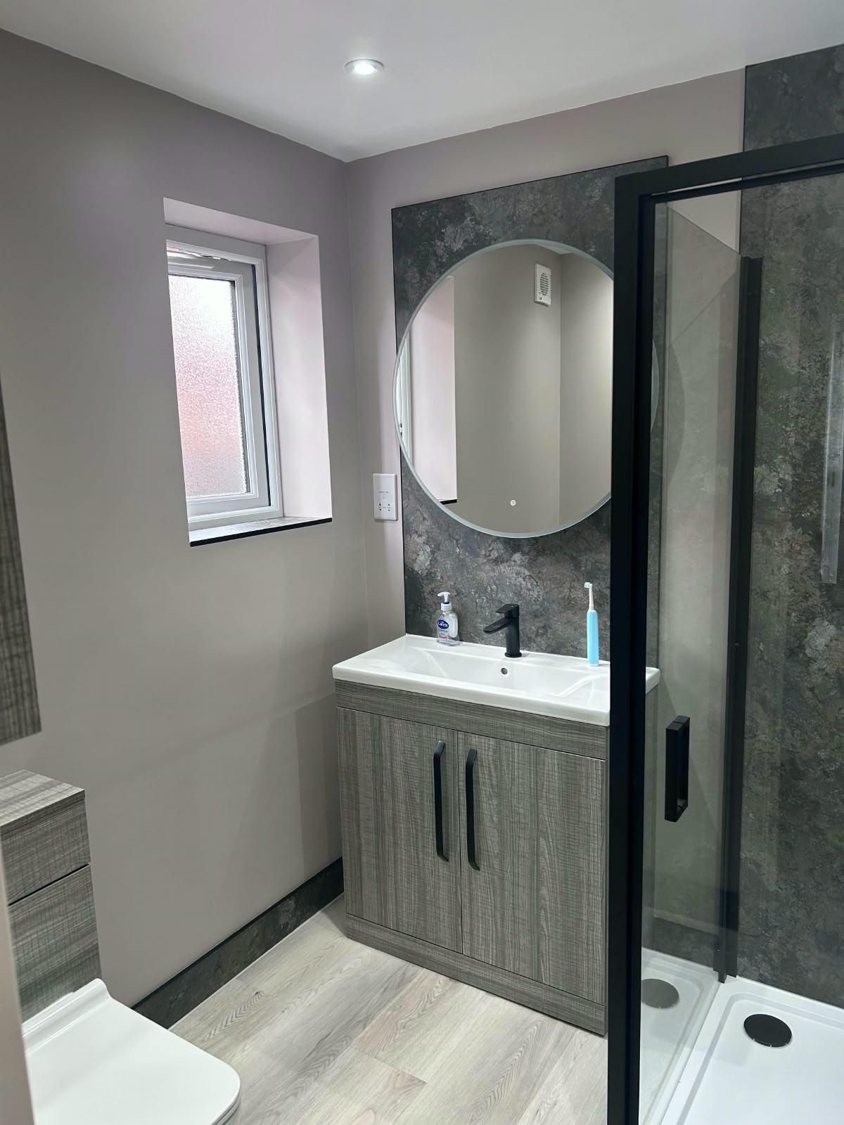 A finished bathroom with a round mirror, grey vanity unit and black-framed shower screen