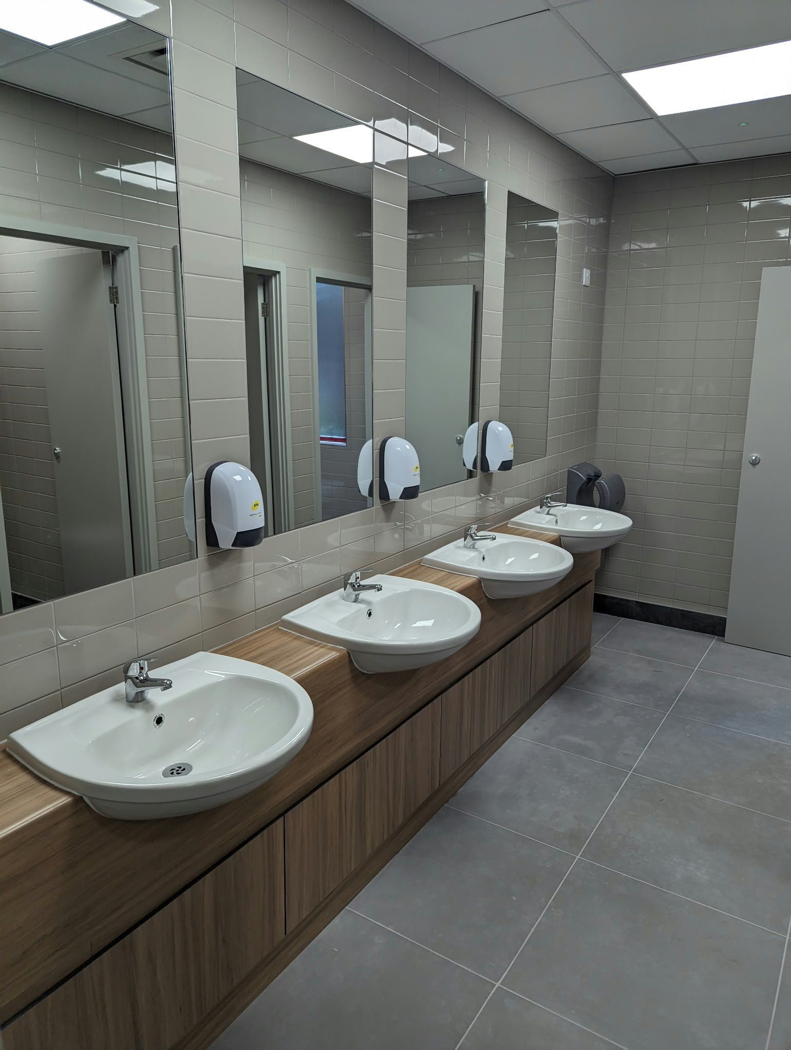 A commercial washroom with three basins set into a wood-effect vanity, with mirrors above