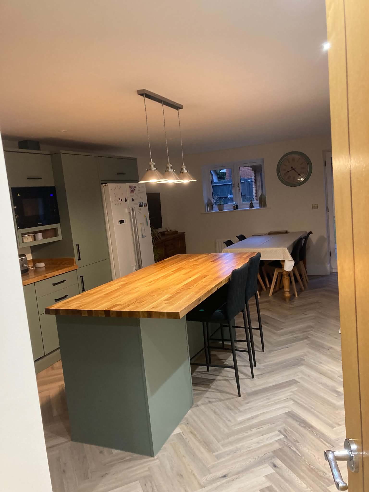 A sage green kitchen with an oak-topped island, pendant lights and herringbone floor