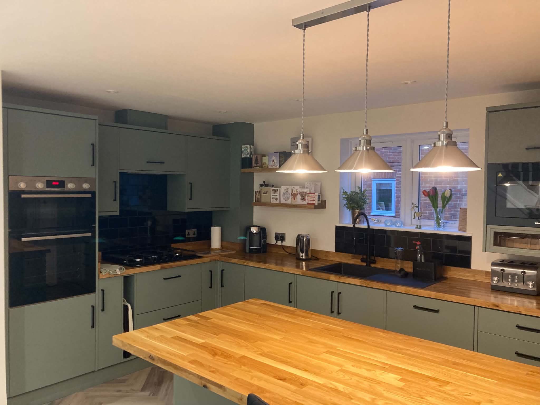 A finished sage green kitchen with oak worktops, built-in ovens and three pendant lights over the island