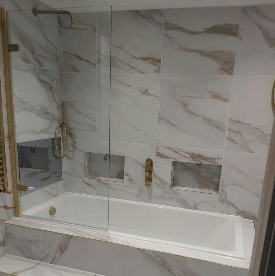 A finished bathroom with marble-effect tiles, a bath with glass screen, brushed gold fittings and two tiled niches